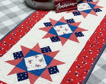May include: A red, white, and blue quilted table runner with a star design. The runner has a white background with red and blue stars and anchors. The text "Proud to be an American" is on a red sign in the background.