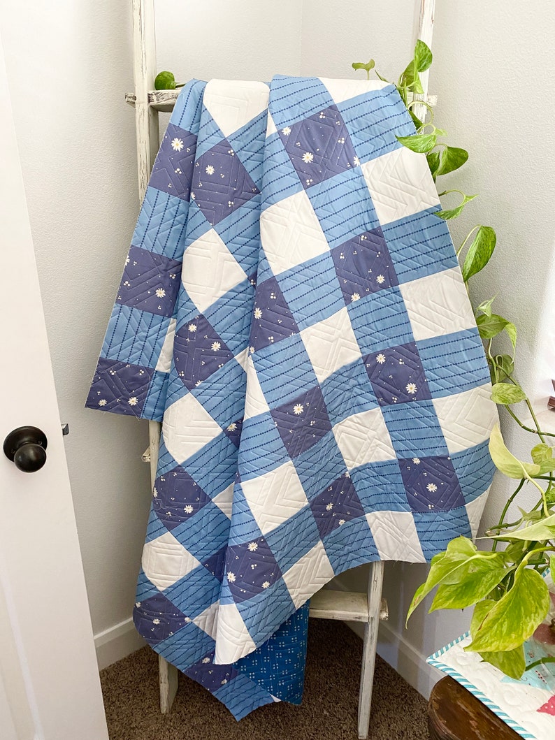 May include: A blue and white quilt with a checkered pattern and a floral design. The quilt is folded and draped over a white wooden ladder.