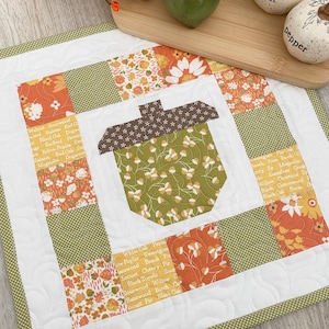 May include: A quilted fabric wall hanging with a patchwork design featuring a large acorn in the centre. The fabric squares are printed with various floral patterns and the names of different types of trees. The fabric is white with a green border.