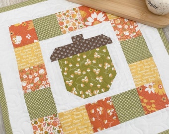 May include: A quilted fabric wall hanging with a patchwork design featuring a large acorn in the center. The fabric squares are printed with various floral patterns and the names of different types of trees. The fabric is white with a green border.