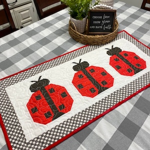 May include: A white table runner with a black and white checkered pattern and a red border. Three red and black ladybugs are appliqued on the runner. The runner is on a white and gray checkered tablecloth. A sign with the text "Choose love, show grace, and have faith" is on the table.
