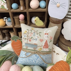 Op de afbeelding: Een decoratieve lentethema-display met een kruissteekkussen met een konijn en kar-ontwerp, omringd door pastelkleurige eieren en een wortel. De afbeelding bevat de tekst "SPRINGTIME STROLL" en "PRIMROSE COTTAGE".
