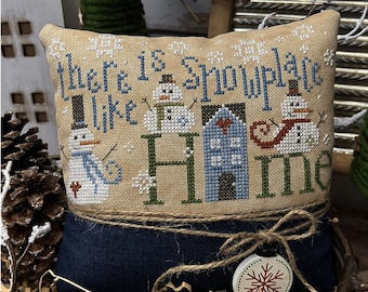 May include: A cross-stitch pillow with the text "There is no snow place like home" and three snowmen. The pillow is on a blue fabric with a brown twine bow. The pillow is in a metal basket with brown paper shreds.