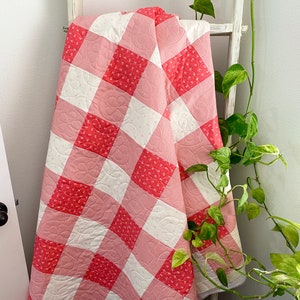 May include: A pink and white gingham quilt with a floral pattern. The quilt is folded and draped over a white ladder.