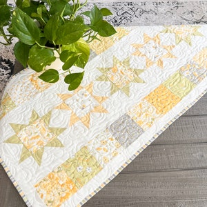 Peut inclure: Un chemin de table blanc avec un motif floral jaune et vert et un design matelassé. Le chemin de table présente un motif d'étoile répété et un bord festonné.