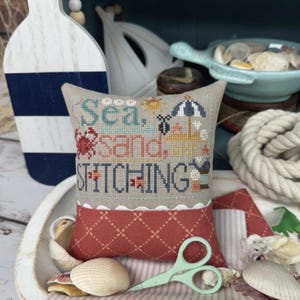 May include: A decorative pillow with the text "Sea, Sand, and Stitching" in cross-stitch. The pillow is red and beige, with a nautical theme. The image includes seashells, scissors, and a wooden boat-shaped decoration.