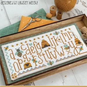 May include: A cross-stitch pattern featuring the alphabet with bees and honeycombs. The pattern is framed in a wooden frame and is titled "A Bee C" and "PRIMROSE COTTAGE STITCHES".