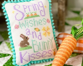 May include: A decorative, square, cross-stitched pillow with the text "Spring Wishes and Bunny Kisses" in purple and green. The pillow has a turquoise pom-pom trim. A woven orange carrot and the text "Spring Wishes" are also visible.