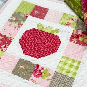 May include: A quilted square featuring a central strawberry design. The strawberry is red with green leaves, surrounded by patchwork squares in pink, green, and floral patterns. The quilt has a white background with a pink border.