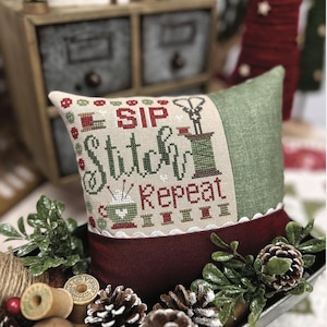May include: A decorative pillow with the text "SIP ♥ STITCH ♥ REPEAT" in red and green cross-stitch. The pillow has a green and burgundy color scheme, with a white scalloped trim. The image includes pinecones, greenery, and spools of thread.