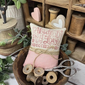 May include: A pink and cream pincushion with the text "Abide In My Love" sits in a wooden bowl with spools of thread and vintage scissors. The pincushion is decorated with a twine bow. The words "Primrose Cottage" are at the bottom.