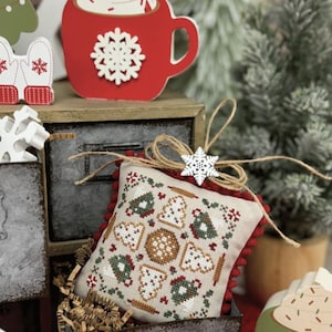 Peut inclure: Une composition de Noël avec une tasse de cacao rouge, un coussin brodé avec des motifs de biscuits, et les textes "Cocoa and Cookies" et "Primrose Cottage". La tasse est ornée de crème fouettée et d'un flocon de neige.