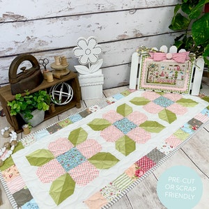 Pode incluir: Um corredor de mesa com um design floral em tons de rosa, verde e azul. O corredor de mesa mede 40,6 cm por 91,4 cm e é feito com tecido pré-cortado ou sobras. O texto "First Blooms Table Runner" e "Primrose Cottage Quilts" está incluído na imagem.