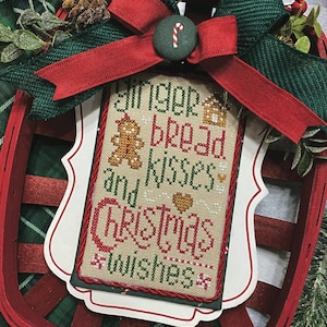 May include: A white cross-stitch sampler with red and green embroidery. The sampler reads "Gingerbread Kisses and Christmas Wishes". The sampler is framed with a white border and is attached to a red and green plaid ribbon.