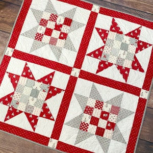 May include: A white and red patchwork quilt with four grey star-shaped blocks. Each block is made of smaller squares of fabric in various shades of grey and white. The quilt is edged with a red border.