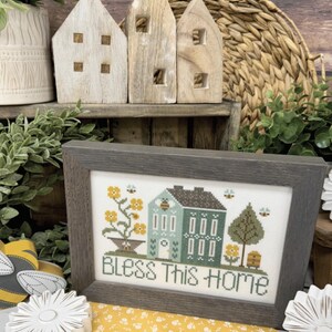以下が含まれることがあります： 「Bless This Home」の文字と家のデザインが施された額入りのクロスステッチ。木製の家の装飾、黄色の花柄の生地、そして「Primrose Cottage」の文字も画像に表示されています。