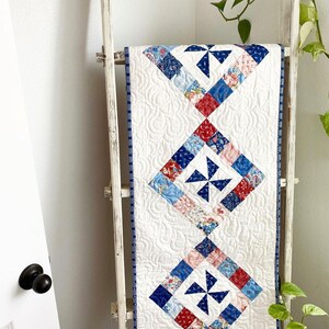 Puede incluir: Un camino de mesa blanco con un diseño de patchwork azul y rojo con molinetes. El camino de mesa tiene un borde azul y blanco y se muestra en una escalera de madera blanca.