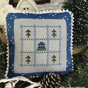 Puede incluir: Una almohada de punto de cruz azul y blanca con una escena de invierno que presenta una pequeña casa, árboles y copos de nieve. La almohada tiene un borde de pompones blancos y está rodeada de ramas de pino y una piña. El texto "January Nine Patch" y "Primrose Cottage Stitches" es visible en la imagen.