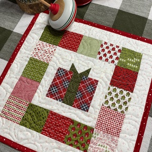 Peut inclure: Un chemin de table matelassé de Noël rouge et vert avec un motif patchwork. Le centre du chemin de table présente un coffret cadeau en plaid rouge et vert avec un nœud vert. Le chemin de table est entouré d'une bordure rouge à pois blancs.
