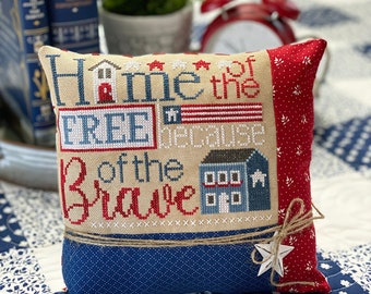 May include: A blue and red pillow with a white background and the text "Home of the Free and of the Brave" embroidered on it. The pillow has a star and a piece of twine tied to it.