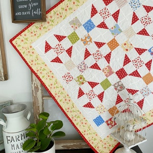 Könnte beinhalten: Eine weiße Steppdecke mit einem roten, weißen und blauen Sternenmuster. Die Steppdecke ist mit einem gelben Blumendruck und einem roten Besatz eingefasst. Die Steppdecke wird auf einem Holzrahmen mit einem Schild präsentiert, auf dem steht "Wähle Liebe, zeige Gnade und habe Glauben."
