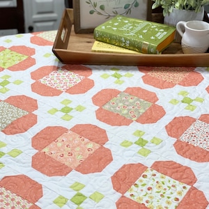 May include: A white quilt with a floral pattern in shades of pink, green, and brown. The quilt is made up of squares and has a quilted design. The quilt is on a table with a wooden tray and books on top.