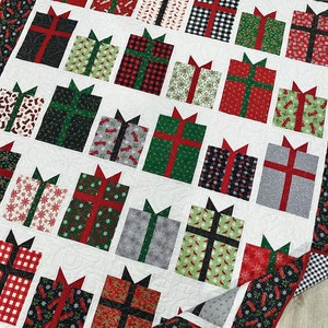May include: A white quilt with a Christmas theme featuring a pattern of red, green, and black gift boxes with various patterns. The quilt is folded in half and the edges are trimmed with a black and white checkered fabric.