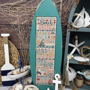 May include: A turquoise surfboard-shaped sign with the text "BEACH RULES" and a cross-stitch design. The cross-stitch reads "Soak up the sun, Ride the waves, Build sandcastles, Roll with the tides, Collect sea shells, Make memories." Decorative nautical elements surround the sign.