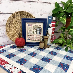 May include: A blue and white gingham quilt with red accents. The quilt is 35 inches by 35 inches and features a small cross-stitch design of a house with a tree and the alphabet. The quilt is on a white wooden surface with a wicker basket, a red ball of yarn, and a potted plant.