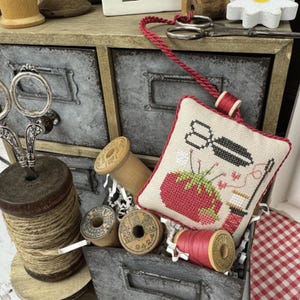 Op de afbeelding: Een rustieke metalen lade-eenheid gevuld met naaiartikelen, waaronder klosjes garen, scharen en een kruissteek scharenfob. De fob heeft een aardbeienontwerp. De tekst "SCISSOR FOB FRENZY", "MARCH" en "PRIMROSE COTTAGE" zijn zichtbaar.