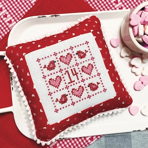 May include: A red and white Valentine's Day pillow with a cross-stitch design featuring hearts, birds, and the number "14". The pillow is on a white tray with a bowl of pink candy hearts.
