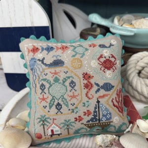 Könnte beinhalten: Ein quadratisches, beigefarbenes Kreuzstichkissen mit einem türkisfarbenen, gewellten Rand. Das Kissen zeigt ein nautisches Design mit Fischen, einem Wal, einer Krabbe, einem Segelboot und den Texten "Beach Quaker" und "Primrose Cottage".