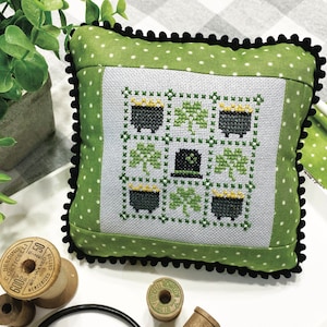 May include: A square decorative pillow with a cross-stitch design featuring pots of gold, shamrocks, and a leprechaun hat. The pillow has a green polka dot border and black trim. Text on the pillow reads "March Nine Patch" and "Primrose Cottage Stitches."