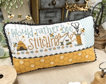Puede incluir: Una almohada de punto de cruz amarilla y blanca con el texto "I Would Rather Bee Stitching". La almohada está decorada con abejas, panales y artículos de costura. La almohada está sentada en una mesa con otros artículos de costura, incluyendo carretes de hilo, tijeras y cuentas.