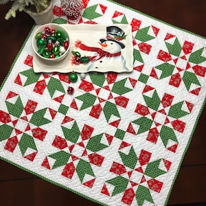 May include: A white table runner with a green and red snowflake pattern. The runner is quilted and has a white border. A snowman figurine is on a white plate on top of the runner.