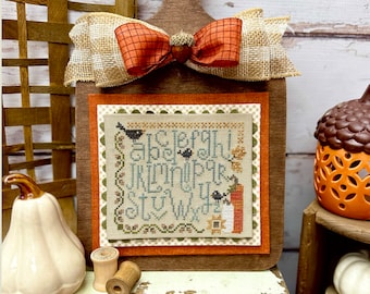May include: A wooden sign with a brown bow and a cross-stitch design featuring the alphabet and the words "Autumn Alphabet" by Lindsey Weight. The sign is displayed on a shelf with other fall-themed decorations.