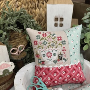 May include: A square pincushion featuring a cross-stitch design of a floral vase, spools, and scissors on a beige background, bordered by light green floral fabric and a red plaid fabric with white flowers, accented by white wooden beads.