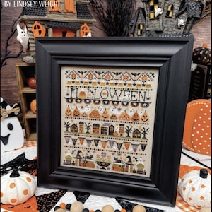May include: A framed cross-stitch sampler titled HALLOWEEN featuring rows of stitched motifs including candy corn, ghosts, jack-o'-lanterns, houses, and black cats on a beige fabric background, enclosed in a simple black wooden frame.