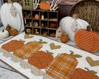 Op de afbeelding: Een herfsttafelloper met oranje geruite pompoenen en hartaccenten. De loper is 84 cm x 43 cm groot en heeft de tekst "Plentiful Pumpkins" en "Primrose Cottage". Decoratieve pompoenen en herfstdecoratie staan op de achtergrond.