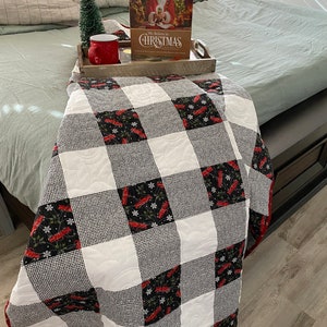 May include: A black and white checkered quilt with a red border. The quilt is folded and placed on a bed with a green bedspread. A book titled "We Believe in Christmas" is on a wooden tray with a red mug and a small artificial Christmas tree.