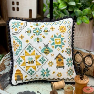Peut inclure: Un coussin blanc avec un motif de point de croix représentant des abeilles, des fleurs et une ruche. Le coussin a une bordure noire avec de petits pompons noirs. Le coussin est posé sur un plateau en bois avec des bobines de fil.