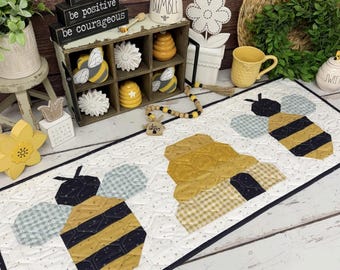 May include: A rectangular fabric table runner measuring 39 inches by 15 inches, featuring a white background with a subtle honeycomb pattern and appliqued designs of two bees and a central beehive in shades of yellow, black, and light blue.