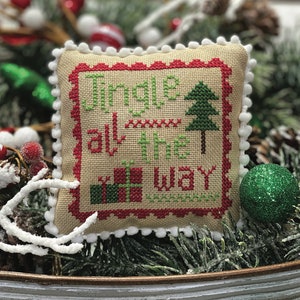 May include: A cross-stitch pillow with a white border and red trim, featuring the text "Jingle all the way" in green lettering. The pillow also includes a green Christmas tree and a red and green gift box. The pillow is sitting on a bed of green pine branches.