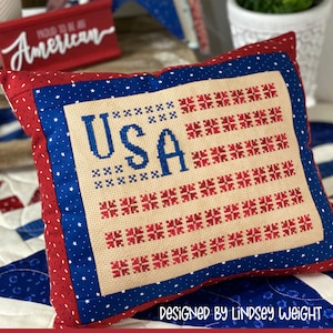 May include: A red, white, and blue pillow with a cross-stitched American flag design and the word "USA" in the center. The pillow is on a blue and white quilt with white stars. The text "Designed by Lindsey Weight Primrose Cottage" is at the bottom of the image.