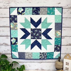 May include: A blue and white quilt with a starburst pattern in the centre. The quilt is made of various fabrics with floral prints and geometric designs. The quilt is hanging on a white wooden wall.