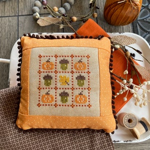 May include: A small orange pillow with a brown trim and a cross-stitch design featuring pumpkins, acorns, and leaves. The pillow is on a white tray with a brown wooden surface in the background.
