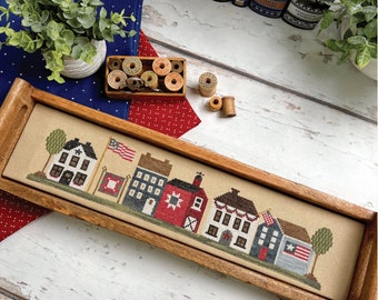 May include: A framed cross-stitch design of a row of houses with red, white, and blue accents. The design is titled "Star Spangled Street" and is by Primrose Cottage Stitches.