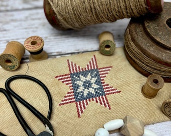 May include: A cross-stitch pattern featuring a red, white, and blue starburst design. The pattern is on a beige fabric and surrounded by wooden spools of thread, scissors, and white beads. The text "flags on the 4th!" and "Release 2" are visible at the top of the image. The text "Primrose Cottage" is visible at the bottom of the image. The text "By Lindsey Weight" is visible in the lower right corner of the image.