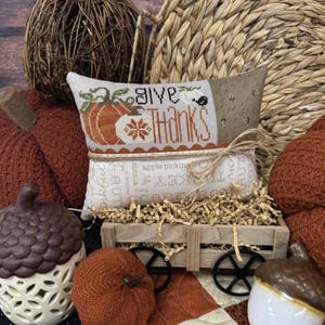 Könnte beinhalten: Eine herbstliche Dekoration mit einem Kissen mit der Aufschrift "Give Thanks" und einem Kürbismotiv. Die Anordnung umfasst dekorative Kürbisse, Eicheln und einen kleinen Holzkarren. Der Text "Primrose Cottage" ist ebenfalls sichtbar.
