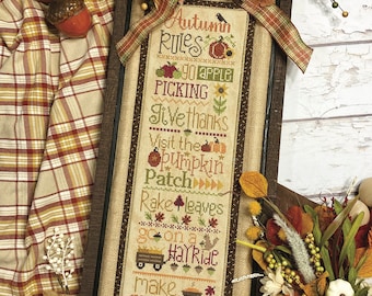 May include: A cross-stitch sampler with a brown wooden frame and a beige background. The sampler features a list of "Autumn Rules" including "Go Apple Picking", "Give Thanks", "Visit the Pumpkin Patch", "Rake Leaves", "Go on a Hayride", and "Make Pumpkin Pie".